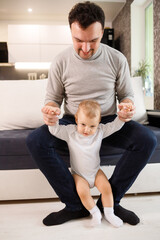 Fototapeta premium Young father assisting walking his infant son teaching to walk at home. Daddy helps son attempting to walk