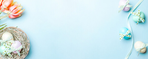 Golden easter colour eggs in basket with spring tulips, white feathers on pastel blue background in Happy Easter decoration. Spring holiday top view banner.