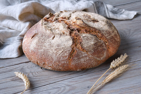 No knead handmade loaf on cutting board on wood with linen towel, spelt ears. German Bauernbrot means Rustic Farmers Bread in English. Wholemeal rye wheat bread baked in Duch oven at home.