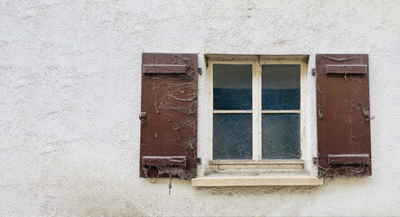 Fototapeta na wymiar Alte Hauswand mit Quadratischem Sprossenfenster, rechts und links braune alte Holzläden und Fenstersims