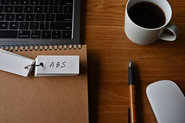 There's a cup of coffee, a laptop and a notebook with a word book on it that says ABS written on...