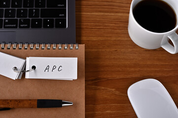 There's a cup of coffee, a laptop and a notebook with a word book on it that says APC written on...