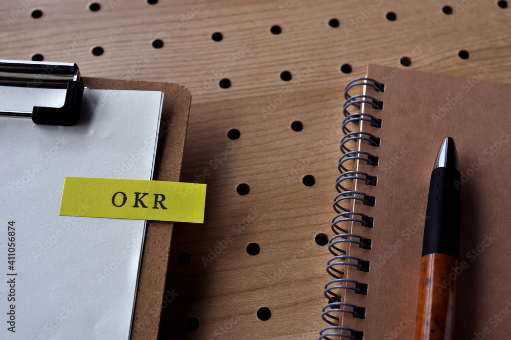 Poster there is a notebook, a pen, and a clipboard with a sticky note stuck to it that says okr written on 