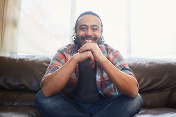 Smiling young latino man in his thirties sitting on his couch at home. - obrazy, fototapety, plakaty