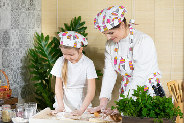 Mom and daughter roll out pizza dough with a wooden rolling pin on one board in the kitchen. Mother and baby do the dough together in the kitchen.