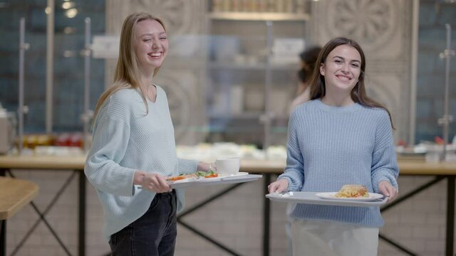 Positive Caucasian and Middle Eastern women posing with food trays in self-service restaurant. Portrait of slim beautiful female friends dining at lunch break in cafeteria. Slow motion.