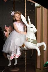 a little girl in a pink dress and a crown on a carousel with a white hare