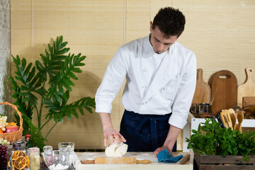 A young handsome chef kneads the dough in the kitchen with one hand on the stool that lies on the...