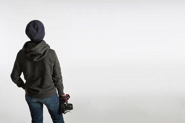 A teenager with a SLR camera on his back stands in a hoodie and a hat on a blue background.