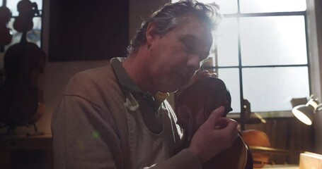 Cinematic shot of experienced master artisan luthier controlling quality of handmade wood violin just made in creative workshop. Concept of spiritual instrument, art, orchestra, passion for music.