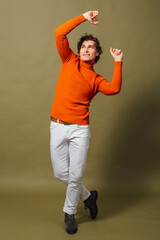 Tall handsome man dressed in orange turtleneck and white jeans posing on the green background