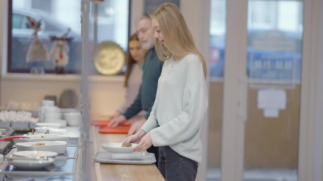 Side view of positive slim young woman ordering dish in cafeteria and leaving queue in slow motion. Smiling beautiful Caucasian lady dining at lunch. Blurred people in line at background.