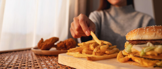 Closeup image of a woman holding and eating french fries and hamburger with fried chicken on the table at home