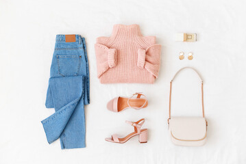 Blue jeans, pink knitted sweater, heeled sandals and small bag with chain strap on white background. Overhead view of woman's casual outfit. Trendy stylish look. Women clothes. Flat lay, top view.
