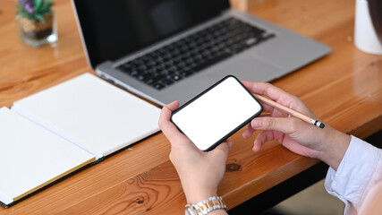 Close up view of young female hands holding horizontal mobile phone while sitting in front of her laptop at office desk.