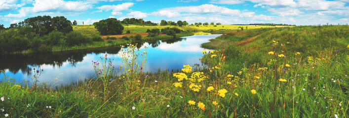 Beautiful summer or spring panoramic rural landscape with calm river and green hills with blooming wild flowers and trees at sunny summer day.