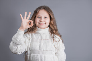 Portrait of a young attractive little girl with blond hair in a white sweater smiling shows okay sign on a gray studio background. Place for text