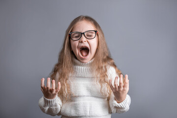 Portrait of a young attractive little girl with blond long flowing hair in a white sweater shouts angrily gestures with hands irritation and screams loudly on a gray studio background.