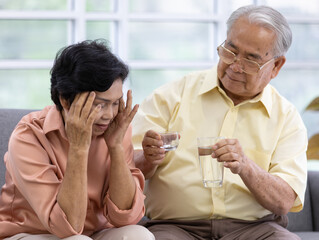 Senior man husband giving a glass of water and cup of medicines for his wife. She is touching her head with her fingers looks like she is in head pain