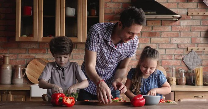 Caring young father teaching small adorable kids cooking in kitchen, enjoying preparing together fresh healthy food. Interested little children siblings learning chopping vegetables with dad.