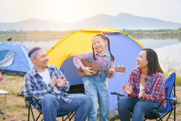 Happy little asian girl playing ukulele and her parents clapping hands at camping site.