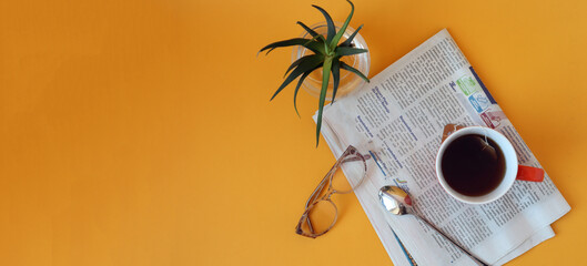 Banner: a cup of tea on a newspaper, glasses for vision, indoor flower alarm clock on a yellow background, top view, space for text