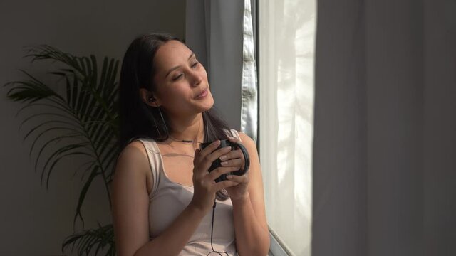 Happy joyful fun smiling woman music lover wearing headphones listening favorite song and crooning while standing near window in morning sunlight.young girl having coffee and enjoying listening music.