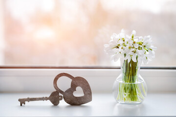 Snowdrops in a vase by the window with a decorative heart-shaped lock. An atmospheric and romantic background for congratulations on Valentine's day. Free space for text