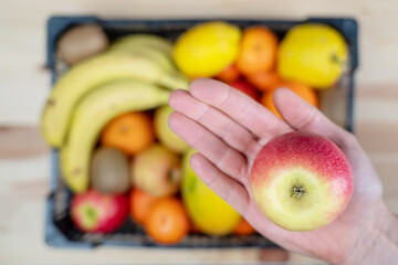 Hand holding an apple in focus and beneath it, is a basket full of varied fruit out of focus.