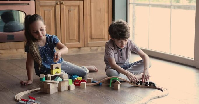 Curious funny little kids brother and sister building wooden towers and riding trains on railroad, enjoying playing together sitting on heated warm floor in kitchen, children domestic hobby activity.