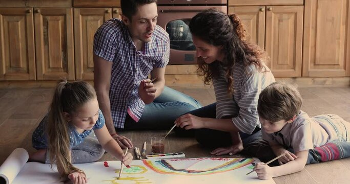 Happy caucasian couple parents enjoying creative domestic hobby activity with little cute son and daughter, drawing pictures with brushes on paper sheet, sitting together on heated floor in kitchen.