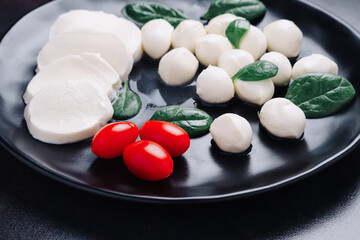 White small mozzarella cheese balls, spinach leaves and tomatoes on black plate.