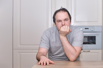 An adult man of 40 years old in casual clothes sits thoughtfully at the kitchen table. Problems in life with despair, grief, and melancholy