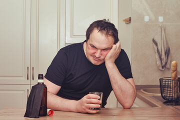 Angry man drinks alcohol sitting with a bottle of whiskey at the kitchen table. Problems with...