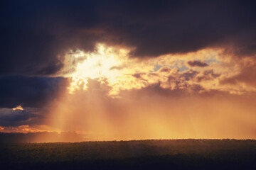Sunbeams make their way through black rain clouds over trees and silhouettes of city buildings
