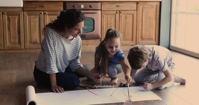 Caring millennial mother sitting on heated warm wooden floor, teaching little cute kids drawing with brushes on poster paper, enjoying creative art domestic family activity together in kitchen.