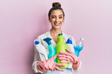 Beautiful brunette young woman wearing cleaner apron holding cleaning products winking looking at...