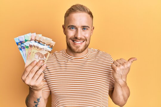 Young caucasian man holding canadian dollars pointing thumb up to the side smiling happy with open mouth