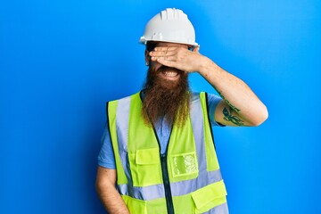 Redhead man with long beard wearing safety helmet and reflective jacket smiling and laughing with hand on face covering eyes for surprise. blind concept.