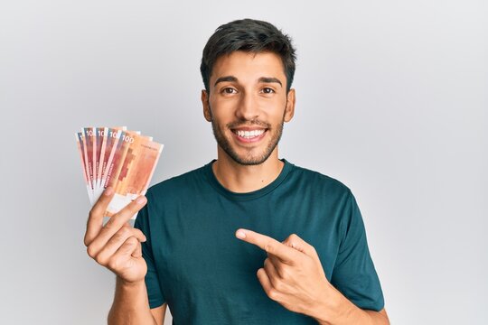 Young handsome man holding 100 norwegian krone banknotes smiling happy pointing with hand and finger