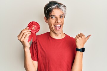 Young hispanic man holding second place badge pointing thumb up to the side smiling happy with open...