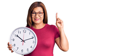 Middle age latin woman holding big clock surprised with an idea or question pointing finger with happy face, number one