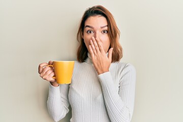 Young caucasian woman holding coffee covering mouth with hand, shocked and afraid for mistake. surprised expression