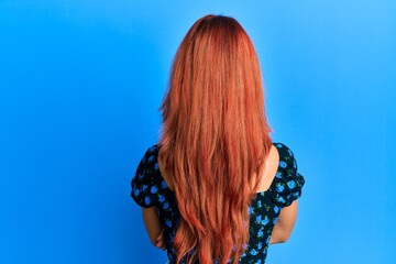 Young beautiful redhead woman wearing casual clothes over blue background standing backwards...