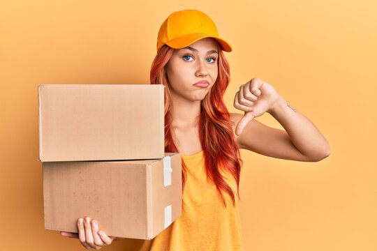 Young redhead woman holding delivery package with angry face, negative sign showing dislike with thumbs down, rejection concept