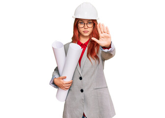 Young redhead architect woman wearing safety helmet holding blueprints with open hand doing stop sign with serious and confident expression, defense gesture