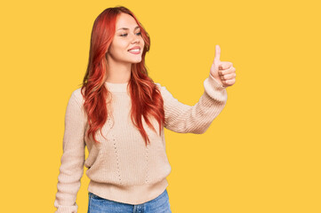 Young redhead woman wearing casual winter sweater looking proud, smiling doing thumbs up gesture to the side