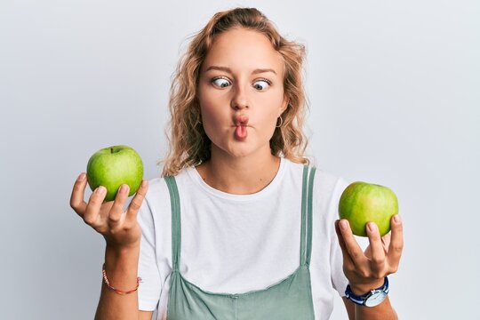 Beautiful caucasian woman holding green apples making fish face with mouth and squinting eyes, crazy and comical.
