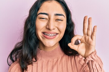 Hispanic teenager girl with dental braces showing orthodontic brackets doing ok sign with fingers,...