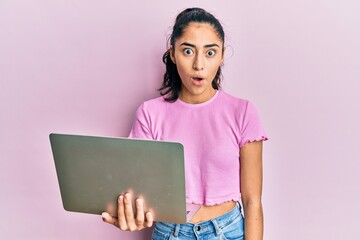 Hispanic teenager girl with dental braces holding and using computer laptop scared and amazed with open mouth for surprise, disbelief face